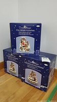 Three boxed polyresin Christmas water fountains by Trim Shoppe stacked, showing overall packaging and festive winter graphics.