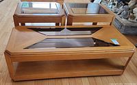Photo showing three wooden coffee tables with glass tops, one large rectangular table with inset geometric glass panels and two smaller square tables with single glass tops.