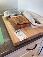Photo showing two wooden cutting boards (one large light wood, one smaller dark wood), grey quilted pot holder, ceramic trivet with black sketch design, and boxed cleaver on countertop.