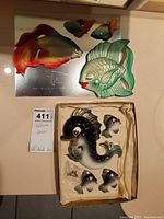Display of all fish wall decorations arranged on a surface. The larger creamy orange-blue fish, green fish, and four black fish pieces shown, one fish with a chip on tail fin.