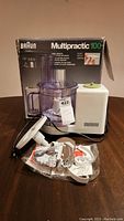 Photo of Braun Multipractic 100 food processor with clear purple bowl, white base, lid, and assorted attachments on a wooden table.