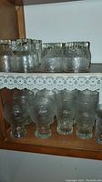 Multiple textured clear glass tumblers, goblets, and small glasses displayed on shelf with lace trim.