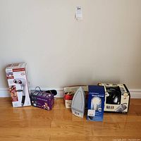 Overview photo showing all four boxed household and personal care appliances including the iron, shower head massager, personal percussion massager, and handheld vacuum.