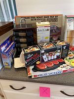 Photo showing assembled lot on kitchen counter with roasting pan set, pasta maker box, dumbbell weights, slicer sets, spice rack, plastic wrap and skewers.