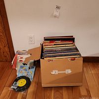 Photo of a cardboard record box filled with 33 and 45 RPM vinyl records near a wall and wooden floor.