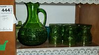 Mid-century green glass pitcher surrounded by six matching green glasses on wooden shelf against white background.