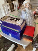 Porcelain doll wrapped in plastic cover next to three boxed sets including a red box, a purple box for the acrylic reindeer set, and another box possibly for nativity items