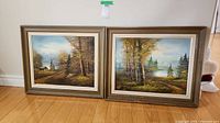 Two framed landscape paintings side by side on floor, showing autumn trees and water.