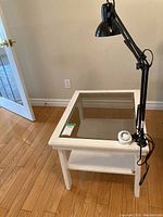 Wood table with glass top, white frame, lower shelf, on hardwood floor, angled view. Black clamp-on lamp attached to table side with round adjustment knob on table top.