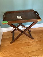 Full view of wooden tray on folding stand placed on hardwood floor against a blue wall. Wooden tray has handles on both ends and shows wear on corners.
