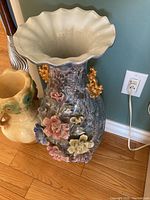 Large decorative vase with multi-colored ceramic flowers, gold ornamental handles, and fluted rim shown against wall outlet