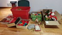 Overview of lot showing large red plastic storage tub, open boxes with colorful glass ball ornaments, vintage figures in box, garland, and tinsel decorations spread on floor