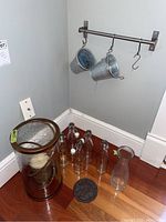 Photo of metal and glass cylindrical candle holder with bronze/copper finish rim and handle, large white pillar candle inside; several clear glass bottles and an 11-inch glass carafe beside it, all placed on floor corner below metal herb rack with 2 hanging galvanized planter pots