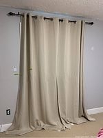 Two beige curtain panels hanging from a dark metal curtain rod with round grommets, showing full length and width of curtains touching the floor.