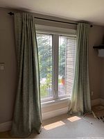 Curtain panels hung on the window showing light green fabric with leaf embroidery and length reaching floor.