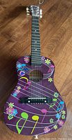 Front view of purple kids acoustic guitar with colorful music note and flower graphics on the body.