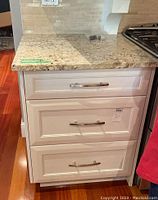 Front view of white lower kitchen cabinet showing three drawer fronts with silver pull hardware and granite countertop.