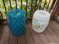 Two ceramic garden stools placed side by side on a wood floor with black metal railing and greenery background, showing overall appearance and relative size.