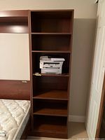 Front view of tall brown wooden bookshelf cabinet with multiple shelves and a printer on one of the shelves. Adjacent to a bed mattress.