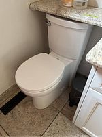 Side view of the white American Standard toilet installed between two surfaces, showing the side flush handle and ceramic finish.