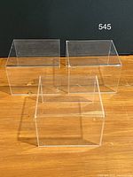 Photo of three clear acrylic display riser units arranged on a wooden surface, showing front and side views. Units are transparent with some visible scratches on surfaces.