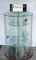 Front view of circular glass display cabinet showing curved glass door with metal hinges and locking knob, four transparent glass circular shelves inside on a central metal rod, and wooden base and rim.