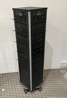 Front and sides view of the black manual rotating display unit with hooks and compartments on all sides, caster wheels visible at base.