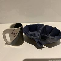 Photo showing the ceramic jug with a handle and geometric black and gray designs next to the deep blue ceramic folded bowl, both artist signed.