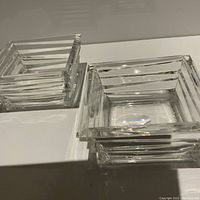 Two square modern crystal bowls with stacked horizontal square layers, displayed on a white surface showing clarity and minor corner chips on the bowls.