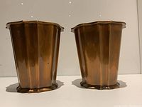 Front angle view of pair of bronze-tone metal planters with scalloped edges on rim and base.