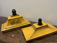 Pair of vintage Italian tole shelf toppers showing yellow enamel surface, black urn handles, decorative black scrolls, and a scene with figures on one piece. Shelf toppers placed on wooden surface with coin for scale.