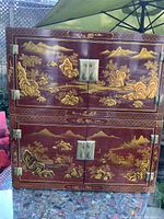 Front view of two stacked lacquer cabinets showing gold painted Asian landscape scenes with mountains and trees, brass hinges and circular plate handles.