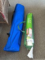 Two bags on carpet floor, one green Coleman labeled beach shade bag, one blue carry bag for folding chair.