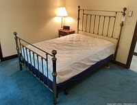 View of brass bed frame from side with mattress and metal legs, showing headboard with vertical spindles and decorative finials in a carpeted bedroom.