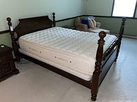 View of dark wooden bed frame with tufted white mattress on cream carpet near window and small furniture.