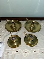 Four brass candle holders arranged on a white embroidered cloth photographed from above. Two larger pieces, two smaller pieces, each with a round drip pan and loop handle.