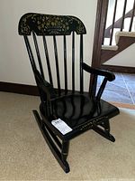 Side angled view of a black wooden child's rocking chair with gold floral and basket accents, showing rocker legs and armrests.