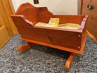 Wooden doll cradle from a side angle showing the rocking legs, nameplate, and bedding inside.