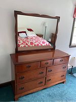 Front view of wooden dresser with mirror showing seven drawers with metal handles, wood grain, polished finish.