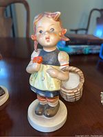 Front view of a Hummel figurine of a girl holding a basket and red object