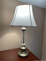 Full view of lamp on wooden surface against beige wall showing glass and metal base and white tapered fabric shade.