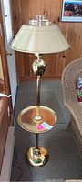 Full view of the floor lamp showing brass base, attached round table, metal lampshade, and glass hurricane shade on top.