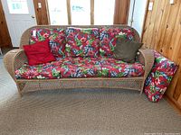 Front view of wicker loveseat with three seat cushions, three back cushions in floral pattern, and two throw pillows (red and olive green).