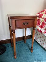 Wooden bedside table shown from a side angle, highlighting the drawer and turned legs, positioned next to a floral bedspread on a blue carpet.