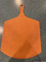 Orange pizza peel with handle and hole for hanging, flat broad paddle with beveled edge, placed on dark countertop.