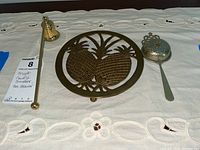 Three items on white embroidered cloth: brass candle snuffer, brass pineapple design trivet, and metal tea strainer with handle.