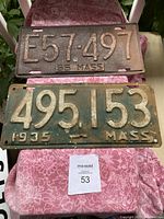 Two antique Massachusetts license plates displayed on a pink patterned cloth. Top plate is red with numbers E57-497 dated 1955. Bottom plate is green with numbers 495-153 dated 1935. Both show heavy rust and wear.