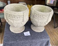 Pair of beige concrete urn-style planters with decorative Greek key and grape cluster relief on a pedestal base shown side by side on a chair.