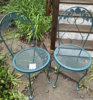 Two green metal garden chairs with decorative backrests placed on outdoor dirt and grass patch near plants.