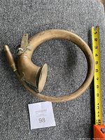 Antique metal car horn showing circular coiled tube with flared bell end and tapered mouthpiece, alongside a measuring tape for scale.